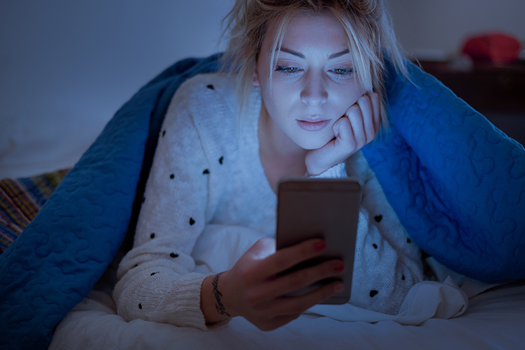 Girl under the sheets reading from her phone at night which prevents a healthy sleep