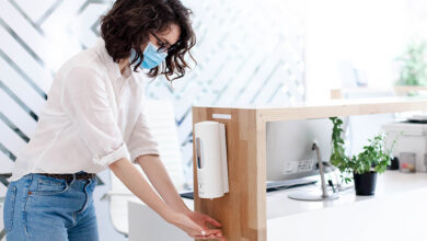 Woman wearing face covering and using automatic hand sanitiser dispenser