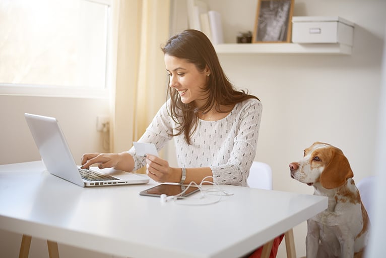 Young woman using laptop in home office with floating shelf behind her and her dog sat next to her