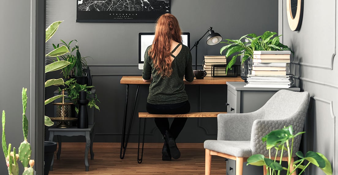 Young woman working on a computer in a home office surrounded by green houseplants.