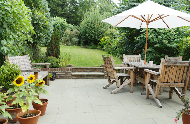 Garden with a paved patio area and seating area with wooden chairs, table and an umbrella, leading up two steps to a lawned area