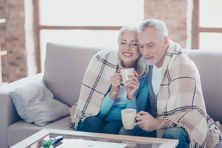 Energy efficiency home grants: Older couple sitting on sofa with blanket and hot drinks