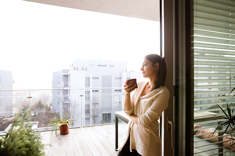 Woman relaxing on apartment balcony with doors open and a hot drink