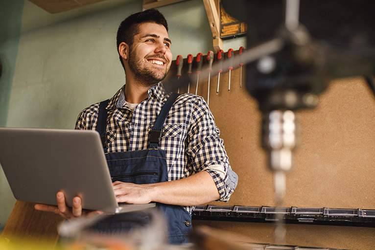 Smiling carpenter in workshop completing business admin on laptop