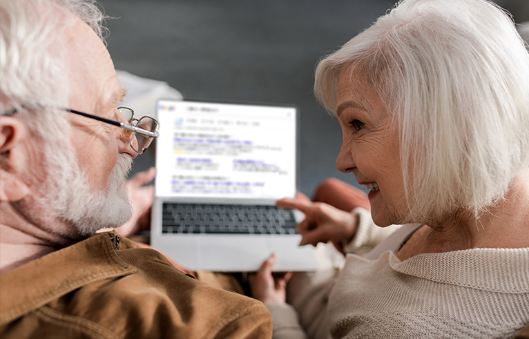 Elderly couple sat facing each other whilst holding an open laptop. 