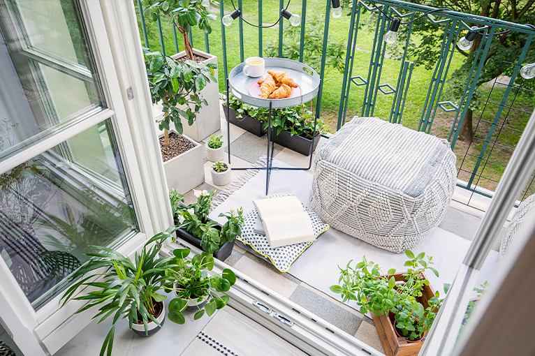 Apartment doors opening out onto a small balcony featuring green plants. a bean-bag and a table with hot pastry and coffee .