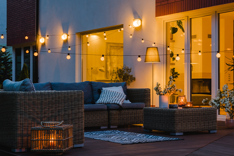 Garden scene at night time alight with low hanging bulbs, a corner lamp and a candle encased in a wooden lantern.
