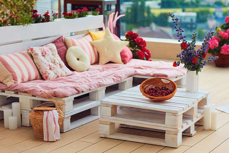 Wooden balcony furniture dressed with pink, red and yellow cushions. Red and purple plants add extra vibrancy to this balcony. 