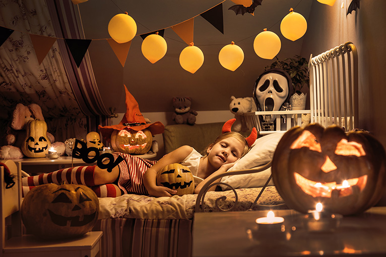 Little girl sleeping in a children's bedroom decorated for Halloween with candle-lit pumpkins. 