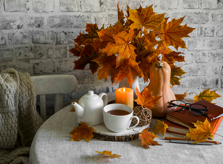 Autumnal table display with a bouquet of orange maple leaves, a tea pot, candle, and brown-leather books.