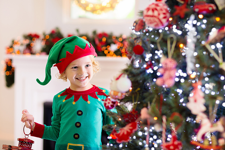 Little boy dressed up as an elf standing by a Christmas tree with the lights switched on. 