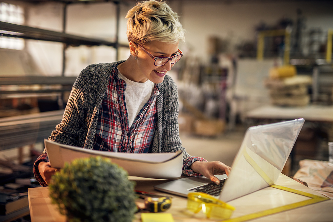 Tradeswoman in a workshop looking at blueprints while typing on her laptop