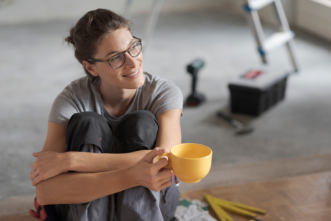 Woman decorator taking a break, sitting on the ground with a cup of tea. Ladder, toolbox, drill and a hammer is visible behind her.