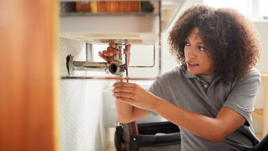 Young black woman plumber sitting on the floor fixing a bathroom sink, seen from doorway