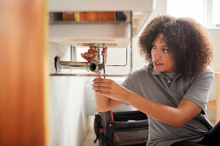 Young black woman plumber sitting on the floor fixing a bathroom sink, seen from doorway