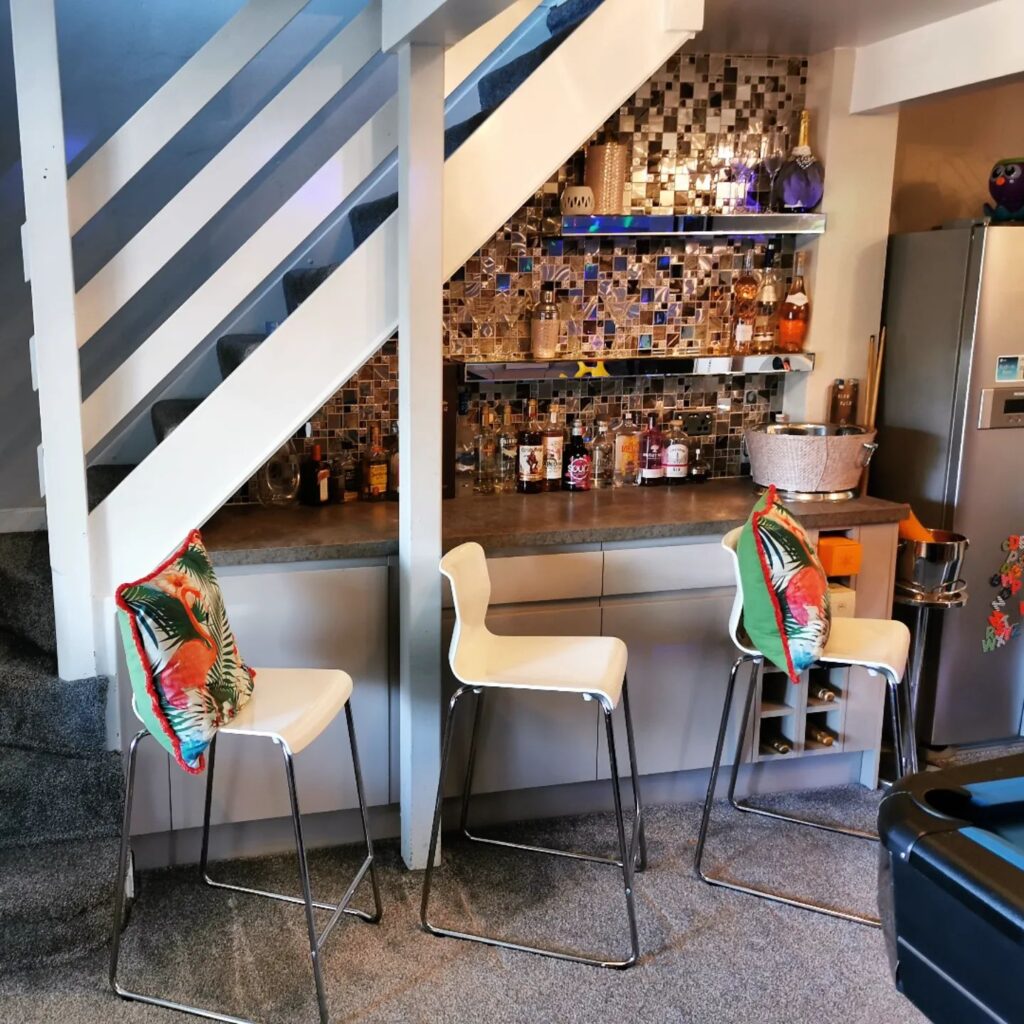 a home bar built under the stairs with three white stools pulled up to it