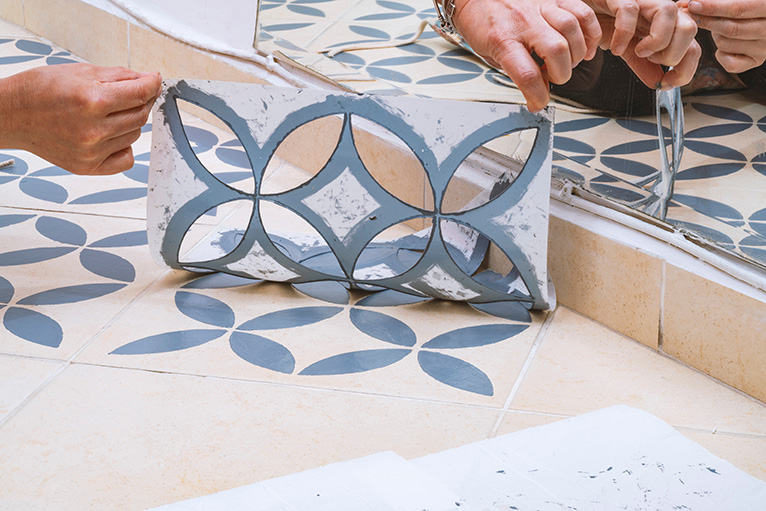Close up of hands removing a decorative painting stencil with a vintage design from the floor tiles after successfully painted into grey.