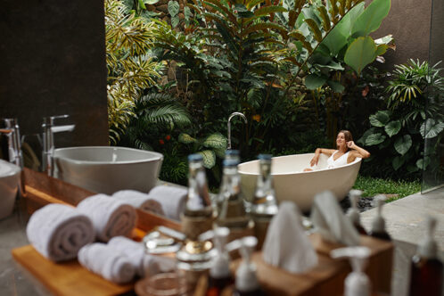 Picture of a bathroom with plants and rolled up towels and a woman in bathtube