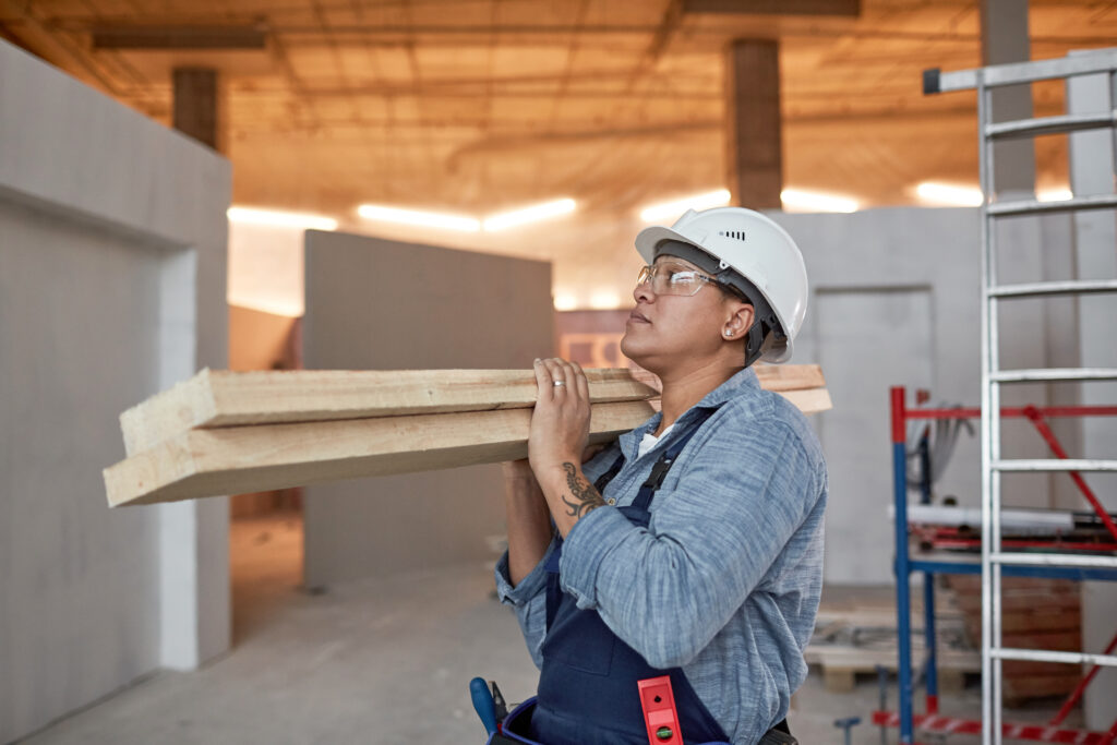 Picture of a female tradesperson in a white hard hat carrying planks of wood